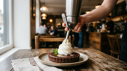 Barista using a stainless steel whipped cream dispenser to pipe a perfect swirl of cream onto a chocolate tart in a warm café setting