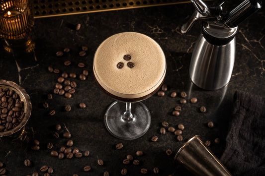 Espresso martini with thick cream foam in a coupe glass next to a stainless steel cream dispenser